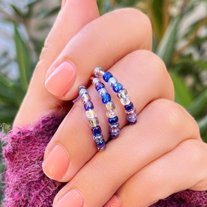 May include: Three beaded rings stacked on a finger. The rings are made of small, colorful beads in shades of blue, purple, and silver.