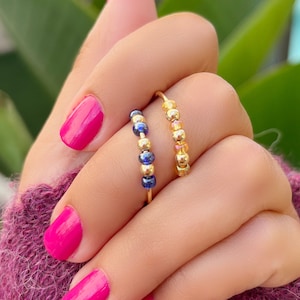 May include: Two gold rings are displayed on a hand with bright pink nail polish. One ring features small blue beads, while the other has gold beads. The background includes green foliage and a purple sweater.