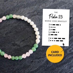 May include: A beaded bracelet with green, pink, and white beads. The bracelet is next to a card that says "THE Psalm 23 MORSE CODE BRACELET" and "CARD INCLUDED". The bracelet is on a dark gray surface.