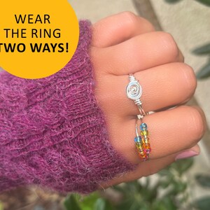 May include: A silver ring with a swirl design is worn on a finger with a rainbow beaded ring. The text "Wear the ring two ways!" is in a yellow circle.
