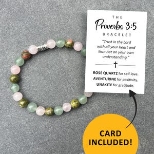 May include: A bracelet made with rose quartz, aventurine, and unakite beads. The bracelet is accompanied by a card with the text "The Proverbs 3:5 Bracelet" and a quote from the Bible. The card also lists the stones and their meanings: Rose Quartz for self-love, Aventurine for positivity, and Unakite for gratitude.