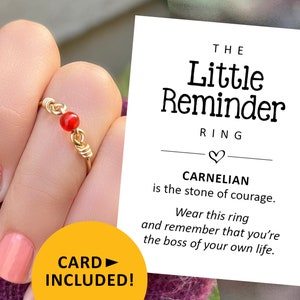 May include: Gold ring with a red carnelian stone. The ring is on a finger and the text "THE Little Reminder RING" is on a white card. The card also says "CARNELIAN is the stone of courage. Wear this ring and remember that you're the boss of your own life." The text "CARD INCLUDED!" is on a yellow rectangle.
