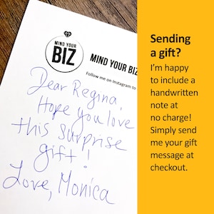 以下が含まれることがあります： 白い紙に青いインクで手書きされたメモ。メモはRegina宛で、Monicaのサイン入り。「MIND YOUR BIZ」のロゴが入っています。画像の右側にある黄色の背景には、ギフトノートに関するテキストがあります。