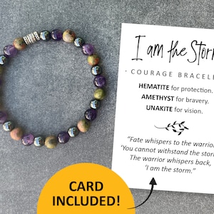 May include: A bracelet made with hematite, amethyst, and unakite beads. The bracelet is accompanied by a card with the text "I am the Storm. Courage Bracelet. Hematite for protection. Amethyst for bravery. Unakite for vision. "Fate whispers to the warrior, 'You cannot withstand the storm.' The warrior whispers back, 'I am the storm.""