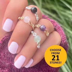 May include: Close-up of four handmade rings with different colored gemstone beads. The rings have silver and gold wire detailing. A yellow circle with the text "CHOOSE FROM 21 STONES!" is in the lower right corner.
