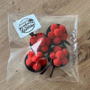 Könnte beinhalten: Klare Plastiktüte mit vier dekorativen Artikeln. Jeder Artikel hat eine schwarze runde Basis mit einem Cluster aus roten Pompons. Sie haben schwarze Stiele. Ein weißes Etikett mit Text ist sichtbar.