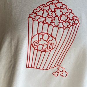 Könnte beinhalten: Ein weißes T-Shirt mit einer roten Umrandung eines Popcorn-Eimers und dem Wort "POPCORN" darin. Es gibt auch rote Umrandungen von Popcorn-Kernen, die um den Eimer herum verstreut sind.