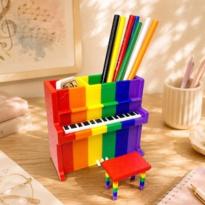 Pode incluir: Um organizador de secretária em forma de piano com as cores do arco-íris, apresentando riscas verticais vermelhas, cor de laranja, amarelas, verdes, azuis e roxas. A frente exibe um teclado preto e branco. Um pequeno banco vermelho a condizer com pernas às riscas arco-íris está colocado à frente.