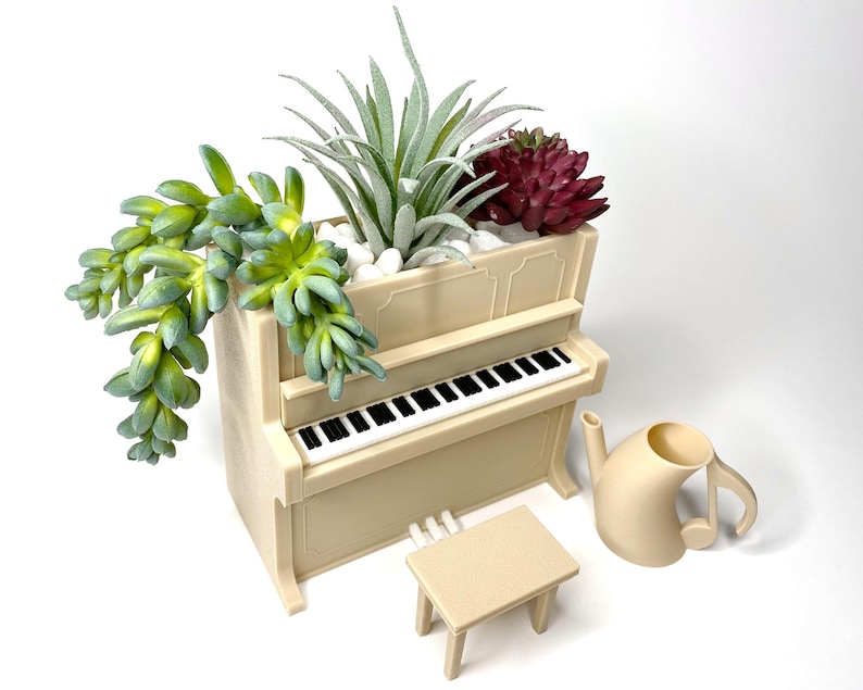 May include: A beige piano-shaped planter filled with succulents and small white stones. A matching stool and a watering can shaped like a musical note sit nearby. The piano has a black and white keyboard.