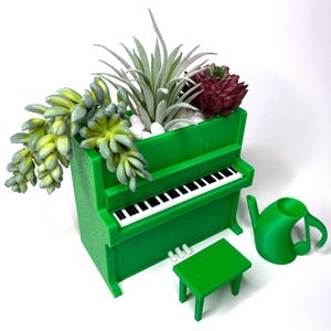 May include: A green piano-shaped planter filled with succulents and small white stones. A miniature green bench and a watering can shaped like a musical note are next to the planter. The piano has black and white keys.