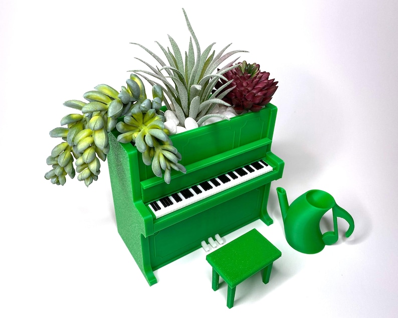 May include: A green piano-shaped planter filled with succulents and small white stones. A miniature green bench and a watering can shaped like a musical note are next to the planter. The piano has black and white keys.