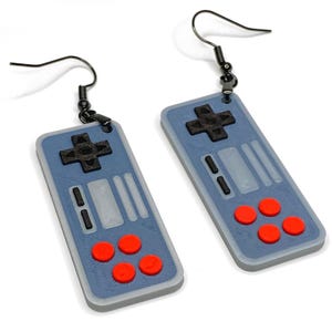Puede incluir: Un par de pendientes con forma de mandos de videojuegos retro. Los pendientes son azules con detalles negros y rojos, incluyendo una cruceta y botones. Los ganchos son negros.