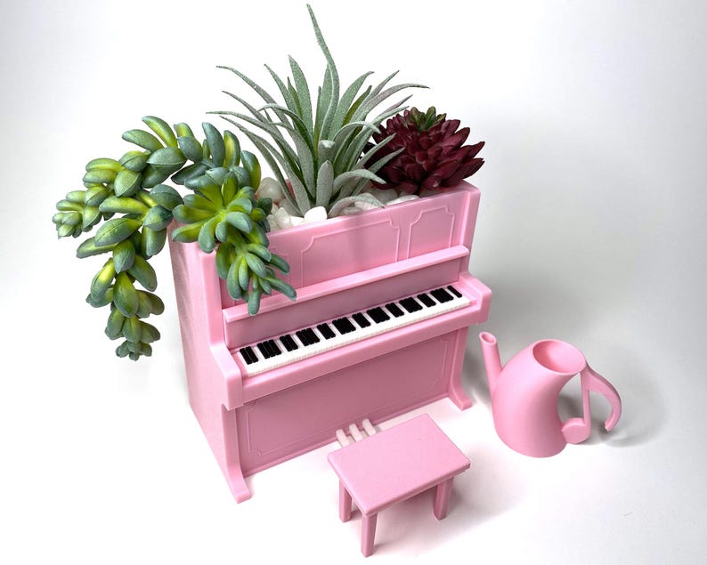 May include: A pink piano-shaped planter with a black and white keyboard, filled with green and burgundy succulents and white pebbles. A matching pink stool and a musical note-shaped watering can are next to the planter.