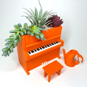 May include: An orange piano-shaped planter with a miniature bench and a musical note-shaped watering can. The planter holds various succulents. The piano has a black and white keyboard. A unique and decorative item for plant lovers.