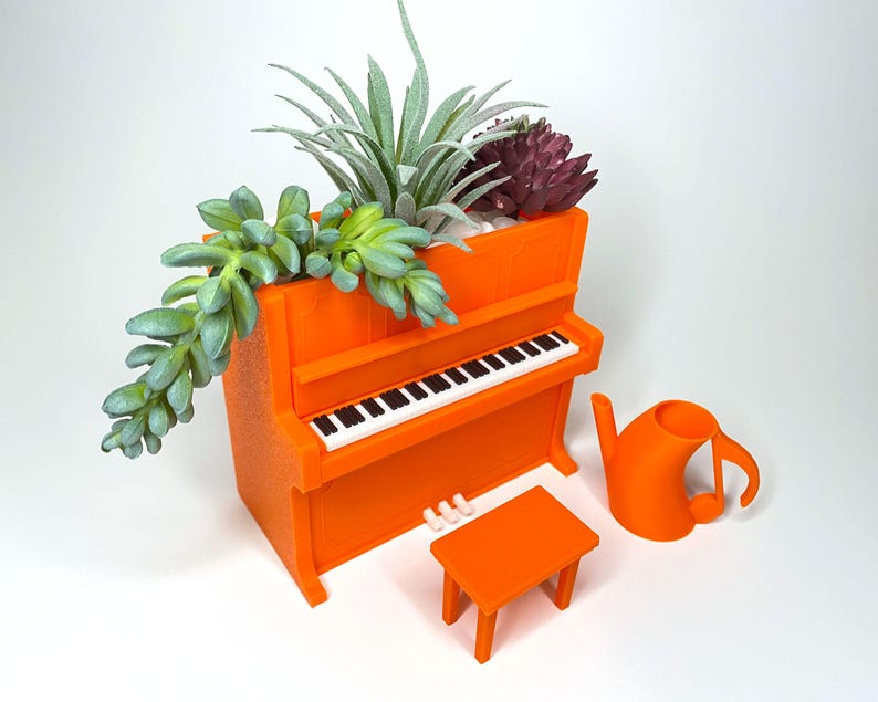 May include: An orange piano-shaped planter with a miniature bench and a musical note-shaped watering can. The planter holds various succulents. The piano has a black and white keyboard. A unique and decorative item for plant lovers.