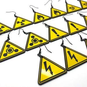Op de afbeelding: Een set van 12 gele driehoekige oorbellen met zwarte waarschuwingssymbolen. De symbolen omvatten een stralingssymbool, een brandsymbool, een uitroepteken, een chemisch symbool, een bliksemflits en een biologisch gevaarssymbool.