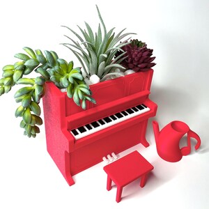 May include: A red piano-shaped planter filled with succulents and small white stones. A matching red stool, piano keys, and a musical note-shaped watering can are also present. The planter is a unique decorative item.