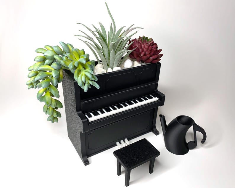 May include: A black miniature piano planter with a white keyboard, holding various succulents and plants. A small black stool and a black watering can shaped like a musical note are next to the piano.
