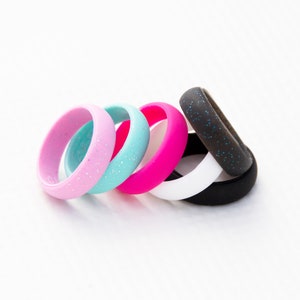 May include: A collection of six silicone rings arranged in a row. The set includes bands in light pink, turquoise, and bright pink, all featuring subtle glitter, alongside a solid white ring, a black ring with blue glitter, and a solid black ring.