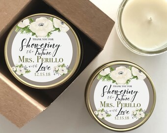 May include: Two small, gold-colored tins with white floral designs and text that reads "Thank you for showering the future Mrs. Perillo with love 12.15.18". The tins contain white candles.