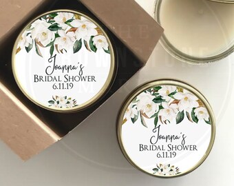 May include: Two small glass jars with gold lids and white labels. The labels feature a watercolor design of white magnolia flowers and green leaves. The text on the labels reads "Joanna's Bridal Shower 6.11.19".