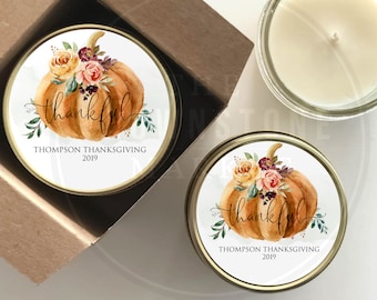 May include: Two small, round, gold-colored tins with white labels. The labels feature a watercolor illustration of a pumpkin with flowers and the text "thankful" in a cursive font. The text "Thompson Thanksgiving 2019" is printed below the illustration. The tins are filled with white candles.