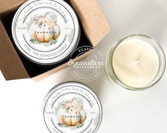 May include: Two small glass jars with lids, each containing a white candle. The lids are decorated with a white and green label featuring a watercolor illustration of a pumpkin with fall leaves and the text "Give thanks to the Lord, for he is good. Psalm 136:1. Thankful. Love, The Johnson Family."