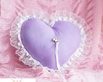 May include: A lavender heart-shaped pillow with white lace trim and a white satin ribbon. The pillow is decorated with a small white flower embellishment.