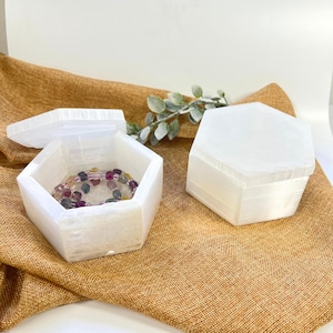 May include: Two white, hexagonal selenite boxes, one open with a colorful beaded bracelet inside, and one closed. The boxes are on a brown, textured surface with a sprig of greenery. The open box's lid is beside it.