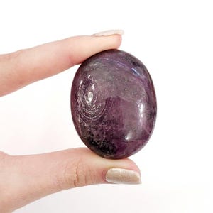 Puede incluir: Una gema púrpura pulida de forma ovalada con patrones arremolinados y un acabado brillante. La piedra se sostiene entre dos dedos sobre un fondo blanco, mostrando su color y textura. La superficie de la gema refleja la luz, resaltando su belleza natural.
