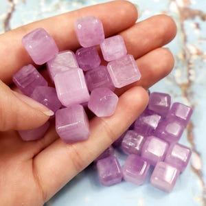 May include: A collection of small, polished, lavender-colored amethyst cubes. The gemstones are held in a hand, showcasing their smooth surfaces and varying shades of purple. The cubes are approximately 0.5 inches in size, with a slightly translucent appearance.