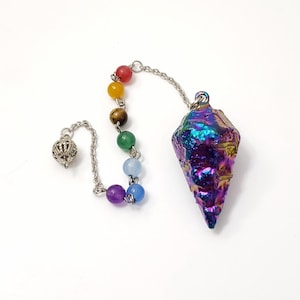 Op de afbeelding: Een pendel met een facetgeslepen, iriserende kristallen punt in paarse, blauwe en groene tinten. Het is verbonden met een ketting met kleurrijke edelsteen kralen en een decoratieve zilveren bedel. De pendel wordt gebruikt voor waarzeggerij.