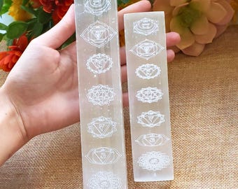 May include: Two rectangular, translucent white selenite wands. Each wand is etched with seven chakra symbols in white. The wands are held in front of a background of flowers and a tan surface.