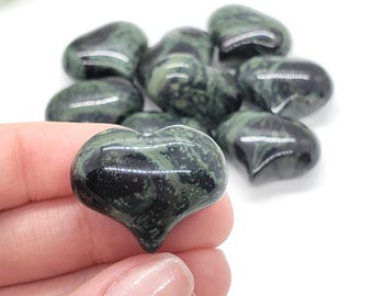 Puede incluir: Colección de piedras pulidas en forma de corazón en tonos verde oscuro y negro. Las piedras tienen un acabado brillante y están dispuestas sobre un fondo blanco. Una piedra en forma de corazón está enfocada, sostenida en una mano.