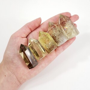 Puede incluir: Un conjunto de cinco puntas de cristal de cuarzo transparente con un tono amarillo dorado. Las puntas están dispuestas en fila, con la punta más grande en la parte superior y la punta más pequeña en la parte inferior.