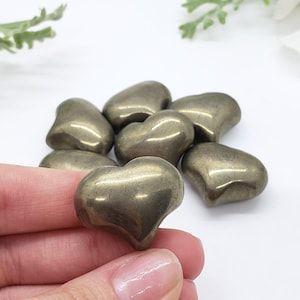 Puede incluir: Un conjunto de piedras de pirita en forma de corazón con un color dorado metálico. Una piedra se sostiene en una mano, con varias otras dispersas en el fondo. Las piedras tienen un aspecto liso y pulido y están dispuestas sobre una superficie blanca.