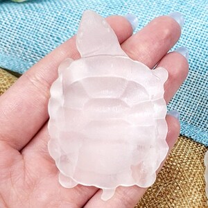 Puede incluir: Una figura de tortuga translúcida de color rosa pálido. La tortuga tiene un patrón de caparazón detallado y pequeños apéndices en forma de aleta. La figura se sostiene en una mano sobre un fondo texturizado azul y beige. Es un artículo decorativo.