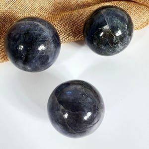 May include: Three polished, dark blue spheres with reflective surfaces. The spheres are arranged on a white surface with a tan burlap cloth in the background. The spheres have a marbled appearance.