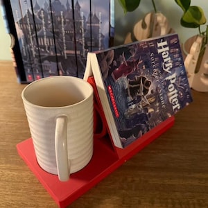 K&ouml;nnte beinhalten: Roter Buchhalter mit einer wei&szlig;en Tasse und einem Exemplar von "Harry Potter und der Stein der Weisen" von J.K. Rowling. Der Buchhalter ist so konzipiert, dass er ein Buch und eine Tasse aufnehmen kann.