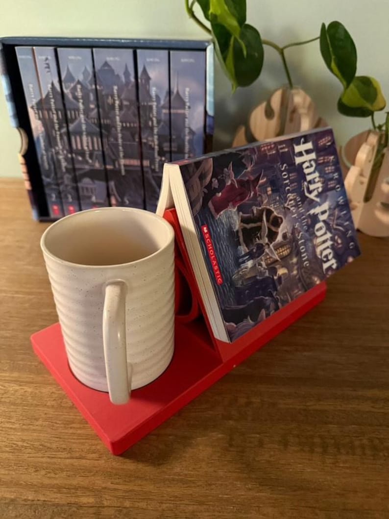K&ouml;nnte beinhalten: Roter Buchhalter mit einer wei&szlig;en Tasse und einem Exemplar von "Harry Potter und der Stein der Weisen" von J.K. Rowling. Der Buchhalter ist so konzipiert, dass er ein Buch und eine Tasse aufnehmen kann.