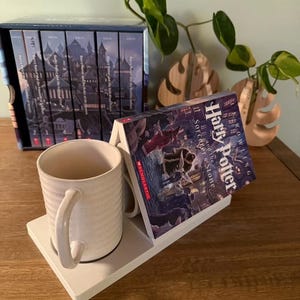 K&ouml;nnte beinhalten: Eine wei&szlig;e Keramiktasse mit gesprenkeltem Finish steht auf einem wei&szlig;en Tassenhalter. Ein Buch mit dem Titel "Harry Potter und der Stein der Weisen" ist auf dem Halter abgest&uuml;tzt. Im Hintergrund befindet sich ein Satz von sieben B&uuml;chern mit einer Schlossillustration auf dem Cover.