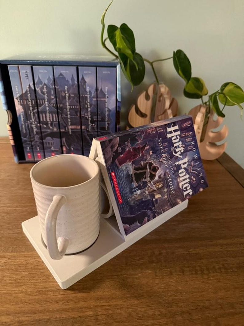K&ouml;nnte beinhalten: Eine wei&szlig;e Keramiktasse mit gesprenkeltem Finish steht auf einem wei&szlig;en Tassenhalter. Ein Buch mit dem Titel "Harry Potter und der Stein der Weisen" ist auf dem Halter abgest&uuml;tzt. Im Hintergrund befindet sich ein Satz von sieben B&uuml;chern mit einer Schlossillustration auf dem Cover.