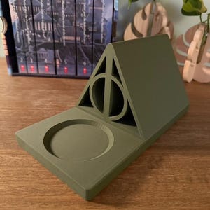 K&ouml;nnte beinhalten: Ein gr&uuml;ner, 3D-gedruckter Harry-Potter-Telefonst&auml;nder mit einem Symbol der Heiligt&uuml;mer des Todes und einer runden Vertiefung f&uuml;r ein Telefon.