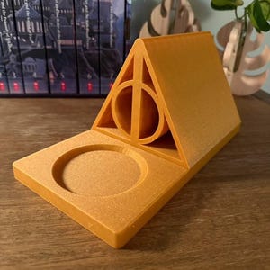 K&ouml;nnte beinhalten: Ein goldfarbener, 3D-gedruckter Telefonst&auml;nder in Form des Symbols der Heiligt&uuml;mer des Todes aus der Harry-Potter-Reihe. Der St&auml;nder hat eine kreisf&ouml;rmige Vertiefung zum Halten eines Telefons oder eines anderen kleinen Gegenstands.