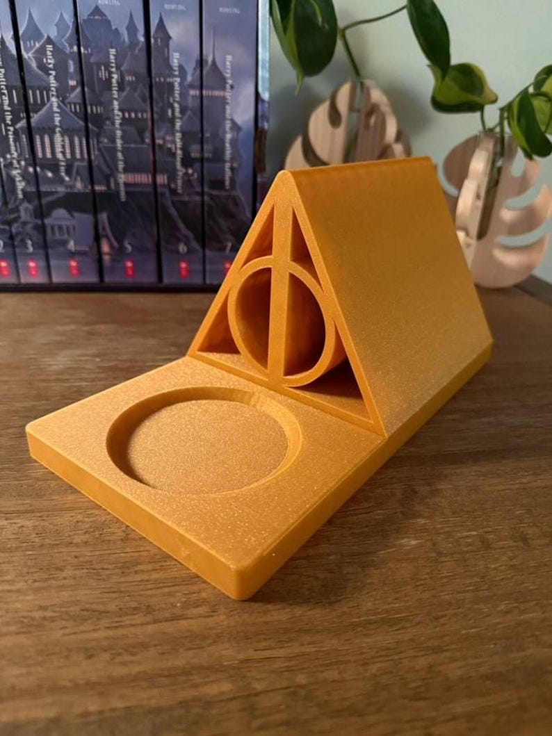 K&ouml;nnte beinhalten: Ein goldfarbener, 3D-gedruckter Telefonst&auml;nder in Form des Symbols der Heiligt&uuml;mer des Todes aus der Harry-Potter-Reihe. Der St&auml;nder hat eine kreisf&ouml;rmige Vertiefung zum Halten eines Telefons oder eines anderen kleinen Gegenstands.