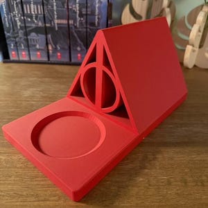 K&ouml;nnte beinhalten: Roter Telefonst&auml;nder aus Kunststoff in Form des Symbols der Heiligt&uuml;mer des Todes aus der Harry-Potter-Reihe. Der St&auml;nder hat eine kreisf&ouml;rmige Vertiefung zum Halten eines Telefons.