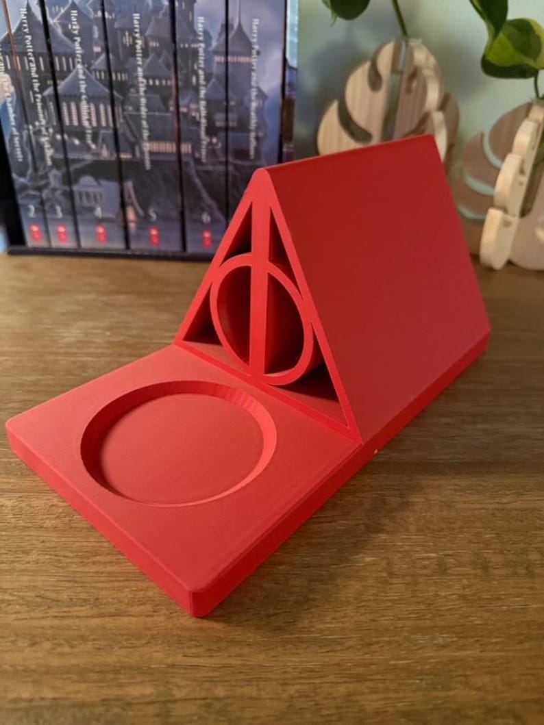 K&ouml;nnte beinhalten: Roter Telefonst&auml;nder aus Kunststoff in Form des Symbols der Heiligt&uuml;mer des Todes aus der Harry-Potter-Reihe. Der St&auml;nder hat eine kreisf&ouml;rmige Vertiefung zum Halten eines Telefons.