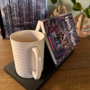 K&ouml;nnte beinhalten: Ein schwarzer Buchst&auml;nder mit einer wei&szlig;en Tasse und einem Exemplar von "Harry Potter und der Stein der Weisen" darauf. Der Buchst&auml;nder steht auf einer Holzoberfl&auml;che.