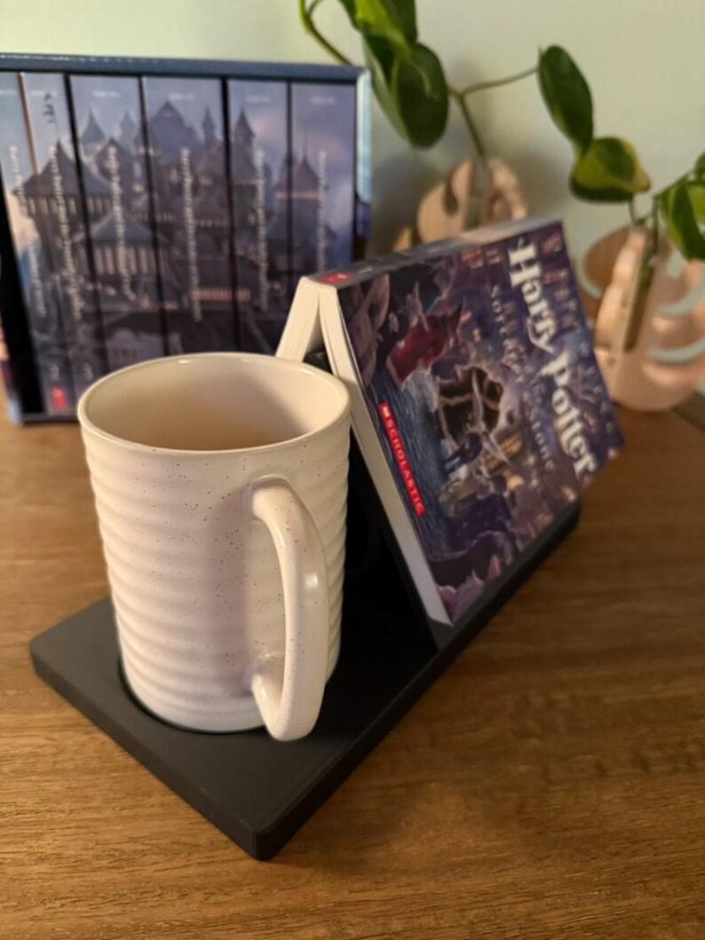 K&ouml;nnte beinhalten: Ein schwarzer Buchst&auml;nder mit einer wei&szlig;en Tasse und einem Exemplar von "Harry Potter und der Stein der Weisen" darauf. Der Buchst&auml;nder steht auf einer Holzoberfl&auml;che.