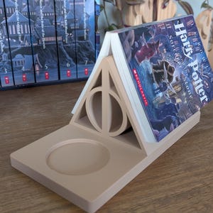 K&ouml;nnte beinhalten: Ein beigefarbener, 3D-gedruckter Buchst&uuml;tze in Form des Harry-Potter-Symbols, den Heiligt&uuml;mern des Todes, mit einem Buch mit dem Titel "Harry Potter und der Stein der Weisen" darauf.